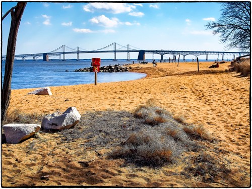 A sandy beach with a bridge in the background, people walking, and a warning sign near the water's edge.