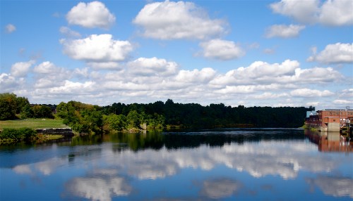 A serene river scene with fluffy clouds reflecting on the water, surrounded by green trees and a brick building.