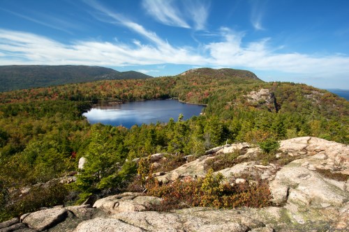 A scenic view of a calm lake surrounded by colorful autumn foliage and rocky terrain under a blue sky with clouds.