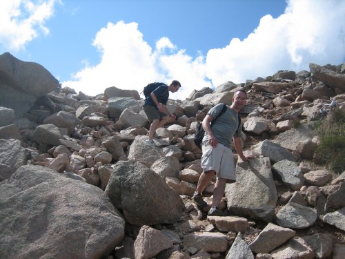 Two hikers navigate a rocky terrain under a blue sky with clouds.