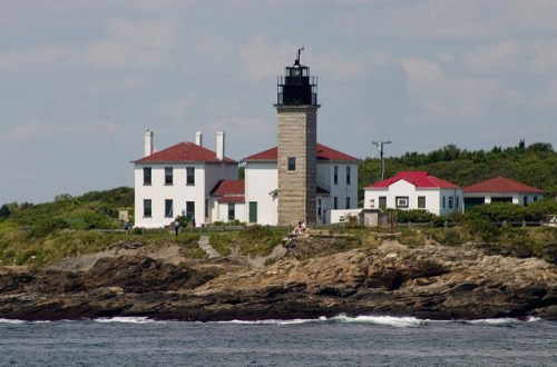 A lighthouse with a red-roofed building stands on rocky shore, surrounded by greenery and ocean waves.
