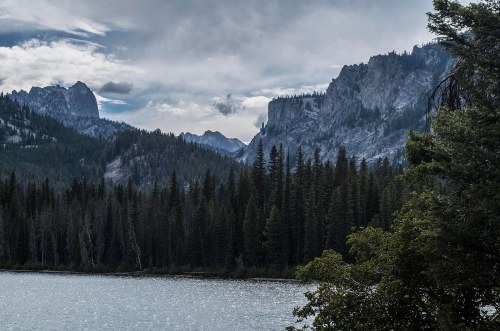 A serene landscape featuring a lake surrounded by dense forests and rugged mountains under a cloudy sky.
