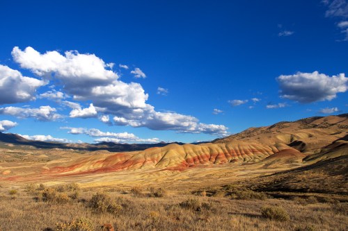 Vibrant, multicolored hills under a blue sky with fluffy clouds, surrounded by dry grassland and distant mountains.