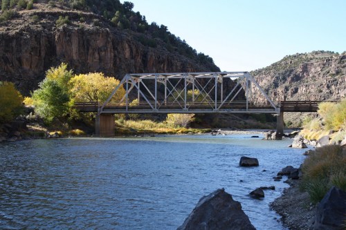 A metal bridge spans a river surrounded by rocky cliffs and autumn-colored trees under a clear blue sky.