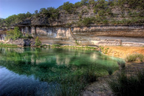 A serene natural landscape featuring a clear green pool surrounded by rocky cliffs and lush vegetation.