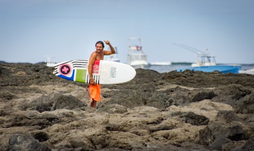 A surfer in orange shorts walks on rocky shore, holding a surfboard and smiling, with boats in the background.