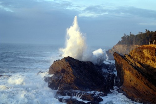 A powerful wave crashes against rocky cliffs, sending a spray of water into the air under a cloudy sky.