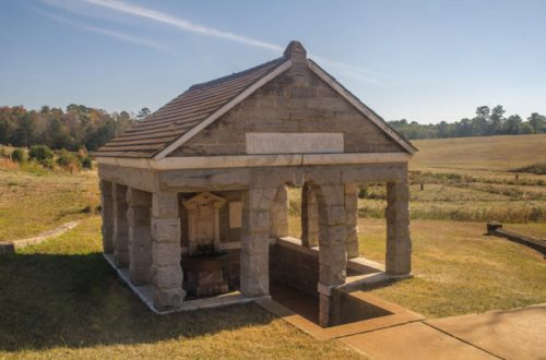A small stone structure with a sloped roof, set in a grassy field under a clear blue sky.