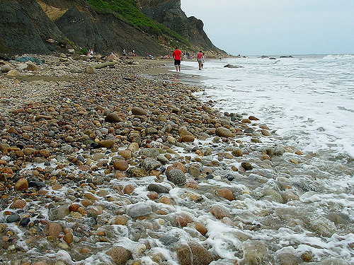 Two people walk along a rocky beach with gentle waves lapping at the shore and cliffs in the background.