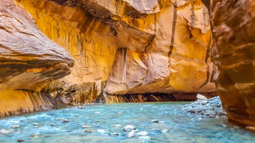 A narrow canyon with vibrant orange walls and a clear blue river flowing through the rocky landscape.