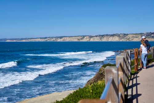 A scenic coastal view with waves crashing against the shore and two people walking along a path by the ocean.