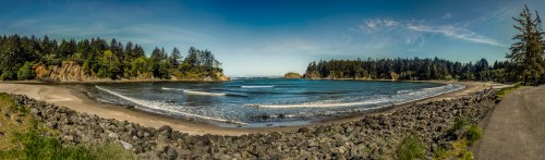 A panoramic view of a serene beach surrounded by trees and rocky shores under a clear blue sky.