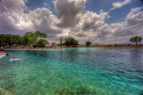 A serene swimming area with clear blue water, surrounded by trees and cloudy skies, with people enjoying the space.