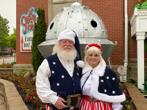 A man in a Santa suit and a woman in a festive outfit pose in front of a silver spaceship and a sign for Castle Noel.