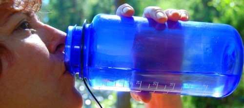 A person drinks from a large blue water bottle, surrounded by greenery in a bright outdoor setting.