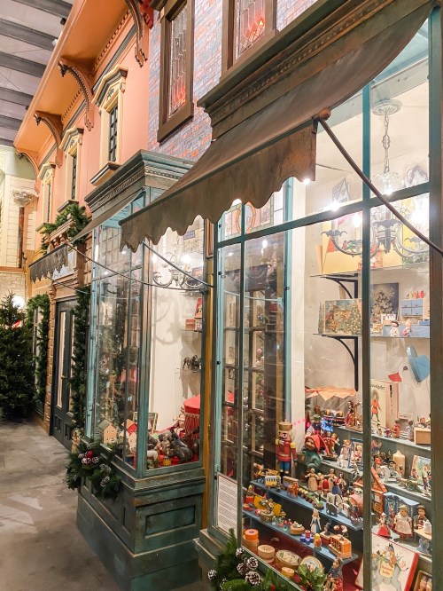 A charming storefront with glass displays filled with colorful toys and decorations, surrounded by festive greenery.