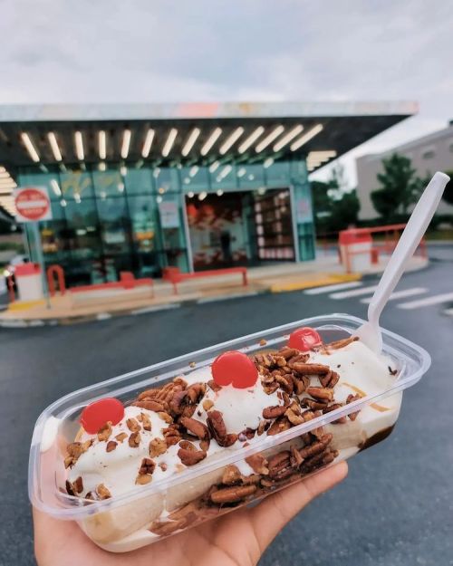 A hand holding a dessert cup with ice cream, nuts, and cherries in front of a modern ice cream shop.