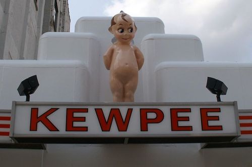 A large, smiling baby figure stands atop a sign that reads "KEWPEE," against a cloudy sky.