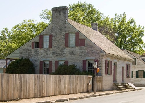 A historic stone house with red shutters, surrounded by trees and a wooden fence, on a sunny day.