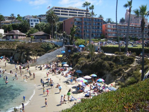 A busy beach scene with people sunbathing, colorful umbrellas, and palm trees, alongside a hotel and rocky cliffs.