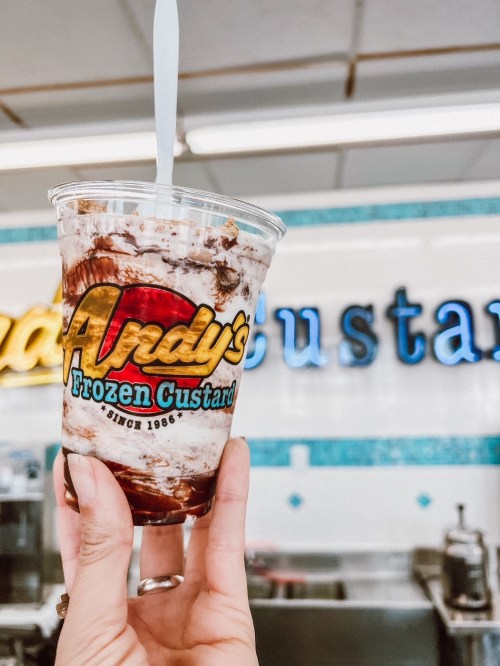 A hand holds a cup of frozen custard with chocolate sauce, in front of a sign for Andy's Frozen Custard.
