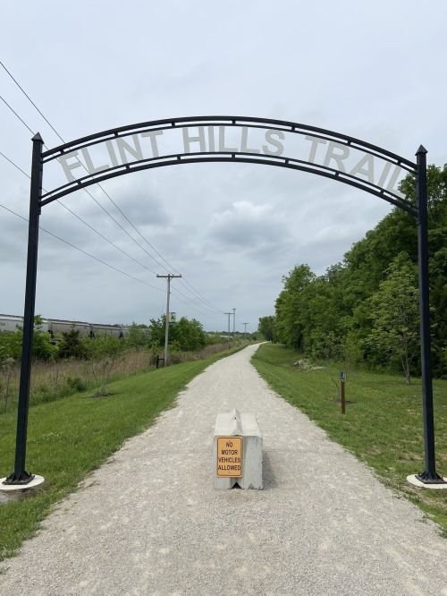 Archway sign for Flint Hills Trail over a gravel path, surrounded by greenery and a warning sign at the entrance.
