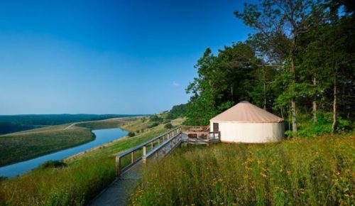 A yurt beside a river, surrounded by lush greenery and rolling hills under a clear blue sky.
