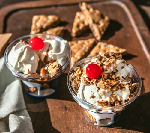 Two dessert cups with whipped cream, nuts, and cherries, alongside pieces of a crunchy dessert on a wooden tray.