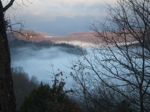 A misty landscape with fog covering a valley, surrounded by trees and distant hills under a cloudy sky.