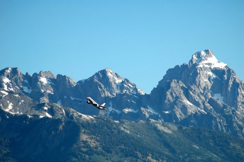 A small airplane ascends against a backdrop of snow-capped mountains under a clear blue sky.