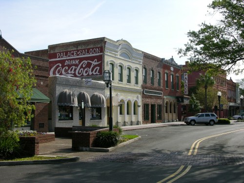Historic buildings line a street, featuring a vintage Coca-Cola sign and charming storefronts under a clear blue sky.
