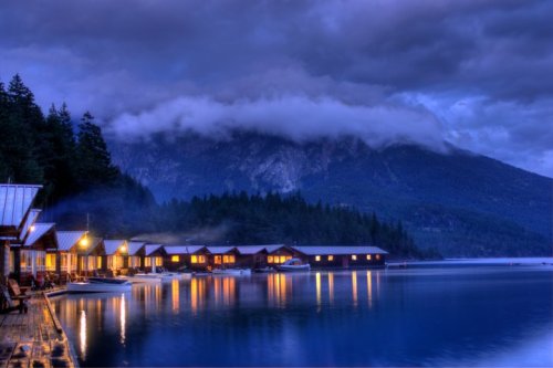 A serene lakeside scene at dusk, featuring cozy cabins with warm lights and mountains shrouded in clouds.