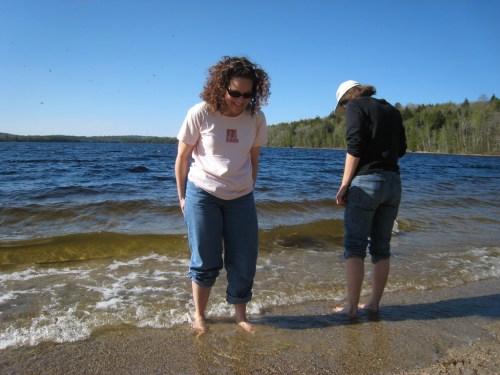 Two women stand at the edge of a lake, dipping their feet in the water on a sunny day. Trees line the background.
