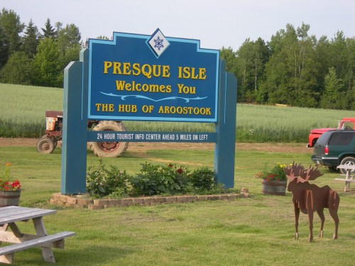 Sign welcoming visitors to Presque Isle, Aroostook, with a moose statue and green fields in the background.