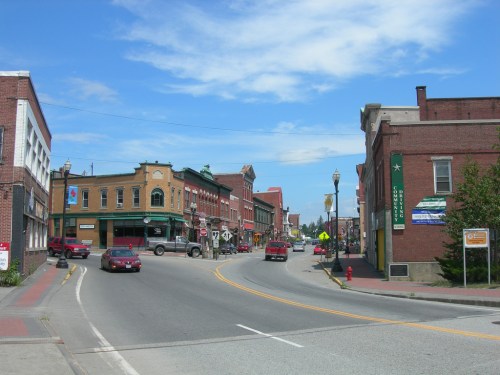A quaint town street with brick buildings, shops, and cars under a blue sky with scattered clouds.