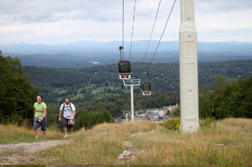 Two hikers walk along a grassy path with a ski lift overhead, surrounded by green hills and a cloudy sky.