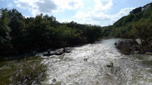 A river flows through a lush green landscape, with two people in a canoe navigating the water.