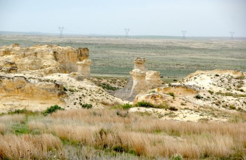 A vast landscape featuring unique rock formations and grassy plains under a cloudy sky. Power lines are visible in the distance.