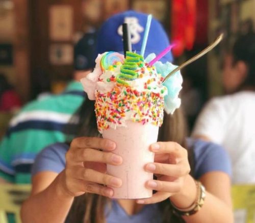 A person holds a colorful milkshake topped with whipped cream, sprinkles, and candy, in a vibrant restaurant setting.