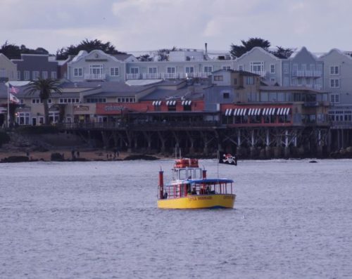 A yellow boat with a pirate flag sails in front of a waterfront building with shops and restaurants.