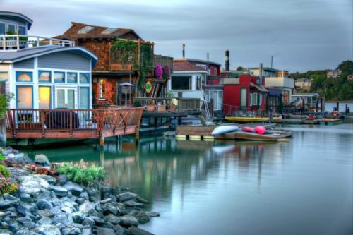 Colorful houseboats lined along a calm waterway, with wooden decks and lush greenery in the foreground.