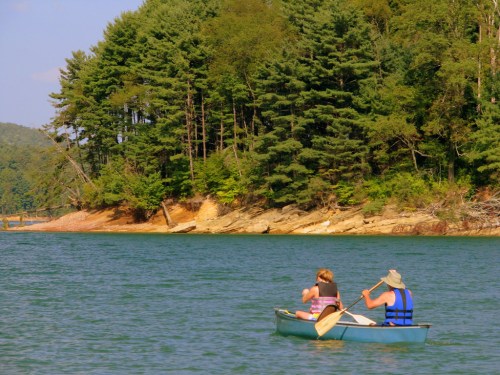 Two people paddle a canoe on a calm lake, surrounded by lush green trees and a rocky shoreline.