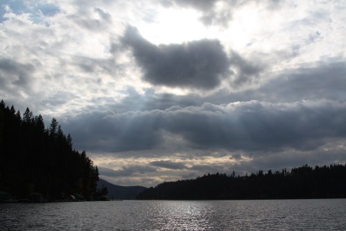A serene lake scene with dark clouds and rays of sunlight breaking through, surrounded by forested hills.