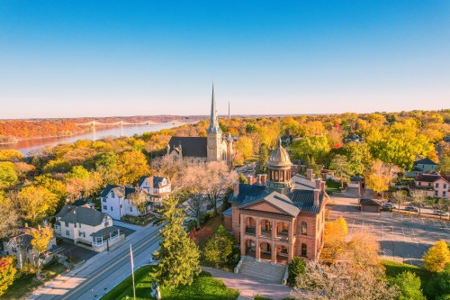 Aerial view of a scenic town with autumn foliage, a church steeple, and a river in the background.