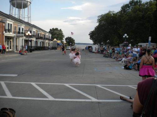 A parade scene with dancers in tutus performing on a street, surrounded by an audience and buildings.