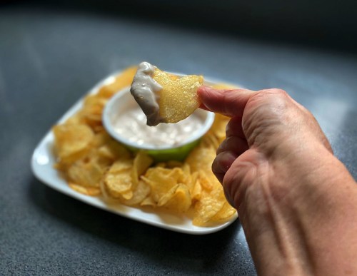 A hand holding a potato chip dipped in creamy sauce, with a plate of chips and a bowl of dip in the background.