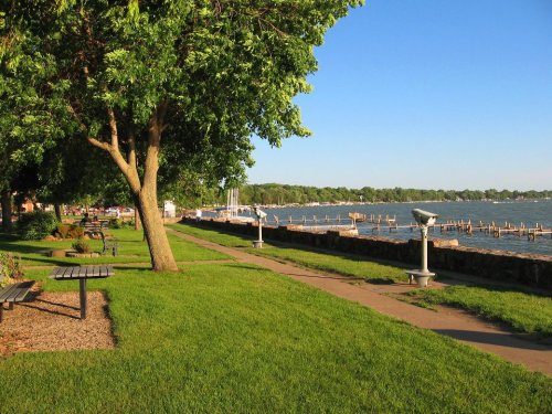 A scenic waterfront park with green grass, trees, benches, and a view of a calm lake and docks under a clear blue sky.