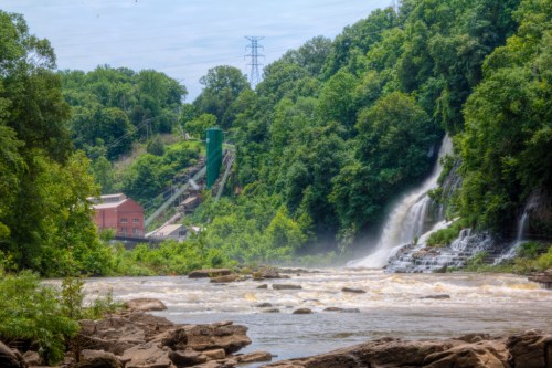 A scenic view of a waterfall surrounded by lush greenery, with a historic building and power lines in the background.