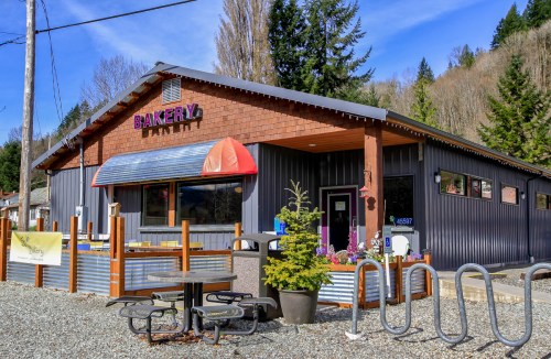 A charming bakery with a colorful sign, outdoor seating, and surrounded by trees on a sunny day.