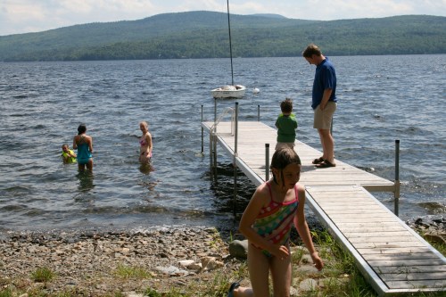 A family enjoys a sunny day by the lake, with children playing in the water and adults on the dock.